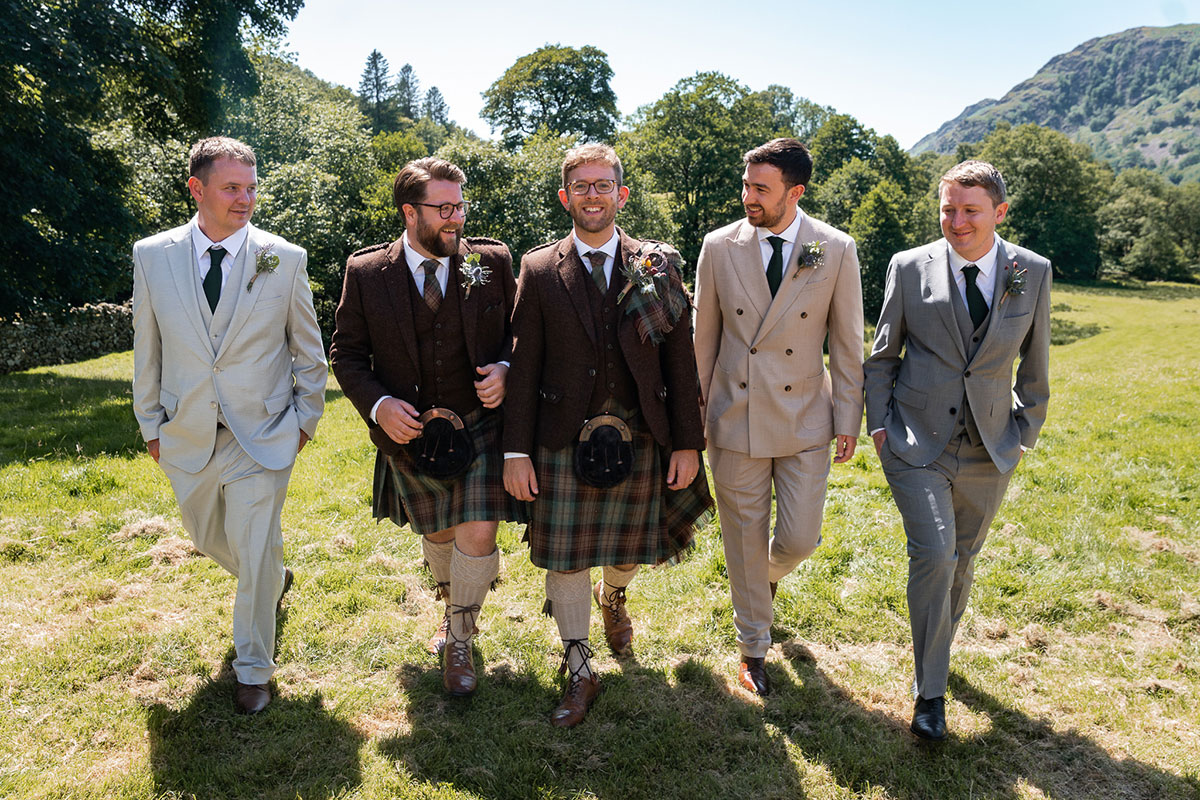 Groom and groomsmen walk together across a grassy field in the sunshine. The groom wears a brown tweed jacket with tartan kilt, while the groomsmen wear a mix of tartan kilts and light-coloured suits