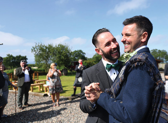 Two grooms wearing Highlandwear holding hands and smiling at each other 