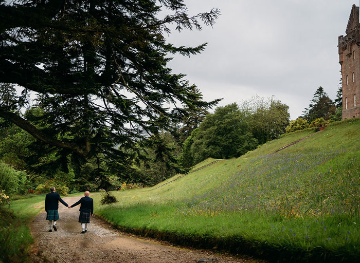 two grooms wearing kilts walk hand in hand along a path next to a grassy slope. There is a large tree on the left and exterior of a castle at the top of the slope on the right