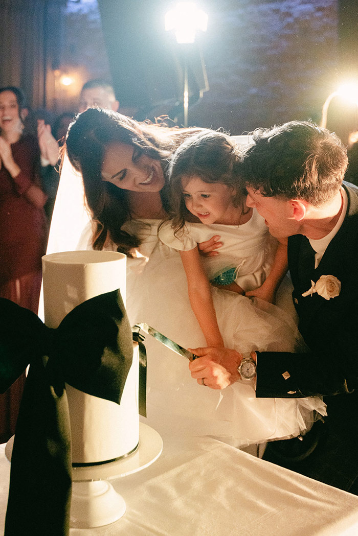 bride and groom hold young flowergirl between them as they help her cut into a white two-tier wedding cake decorated with a large black bow