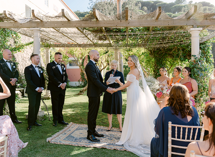 a bride and groom stand on a rug under a wooden structure in a garden with bridesmaids and men in tuxedos on either side and seated wedding guests looking on