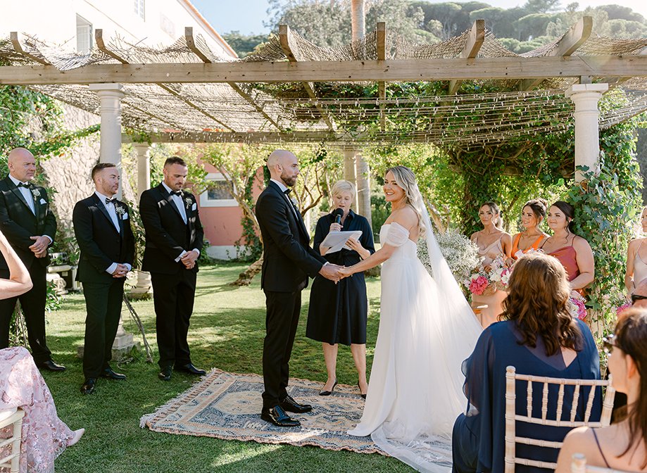 a bride and groom stand on a rug under a wooden structure in a garden with bridesmaids and men in tuxedos on either side and seated wedding guests looking on