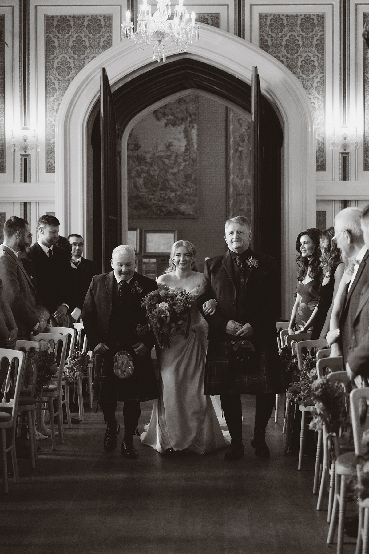 Bride walking down aisle with family during emotional indoor Scottish wedding ceremony in elegant venue