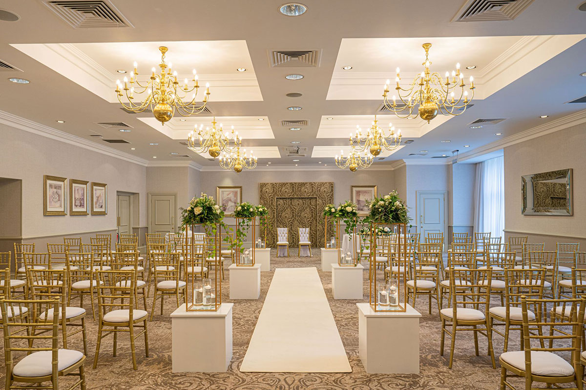 indoor ceremony set-up on patterned brown swirly carpet with golden chairs in rows and tall floral aisle statement pieces lined up along the side of the aisle runner