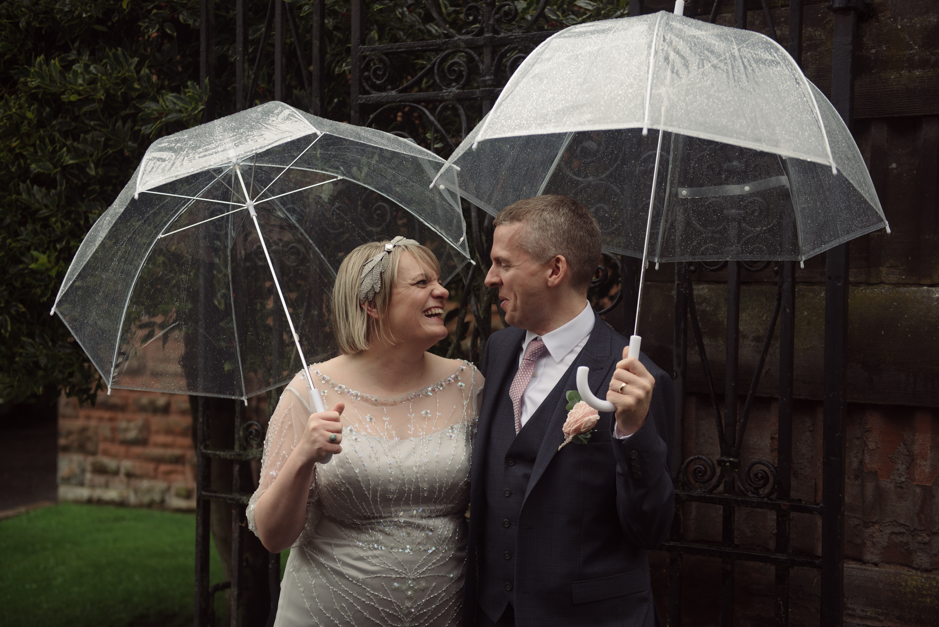 couple smiling wearing wedding outfits as the each hold an umbrella in the rain
