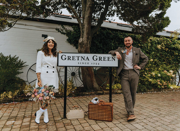 a bride and groom standing either side of a road sign that reads 'Gretna Green'. The bride is wearing a white blazer, a white beret and white boots and carrying a large pastel coloured bouquet. The groom is wearing a brown tweed suit with orange handkerchief tied around his neck