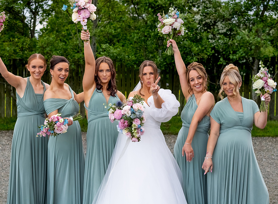 Six women, one wearing white and five wearing teal dresses, posing for the camera whilst holding colourful bouquets 