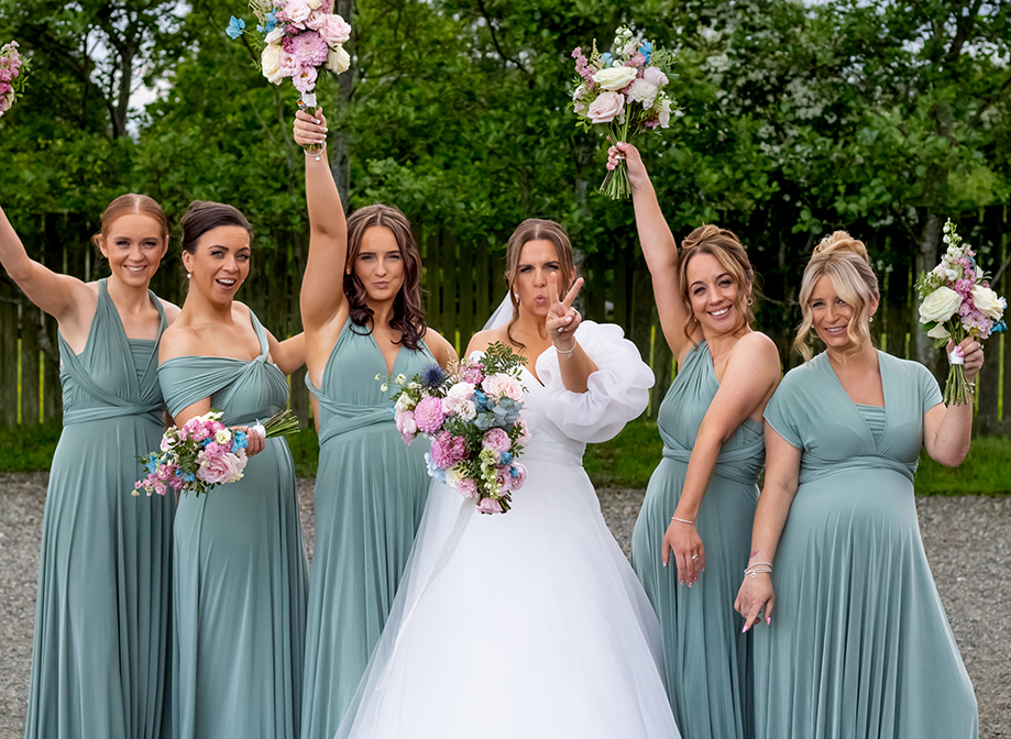 Six women, one wearing white and five wearing teal dresses, posing for the camera whilst holding colourful bouquets