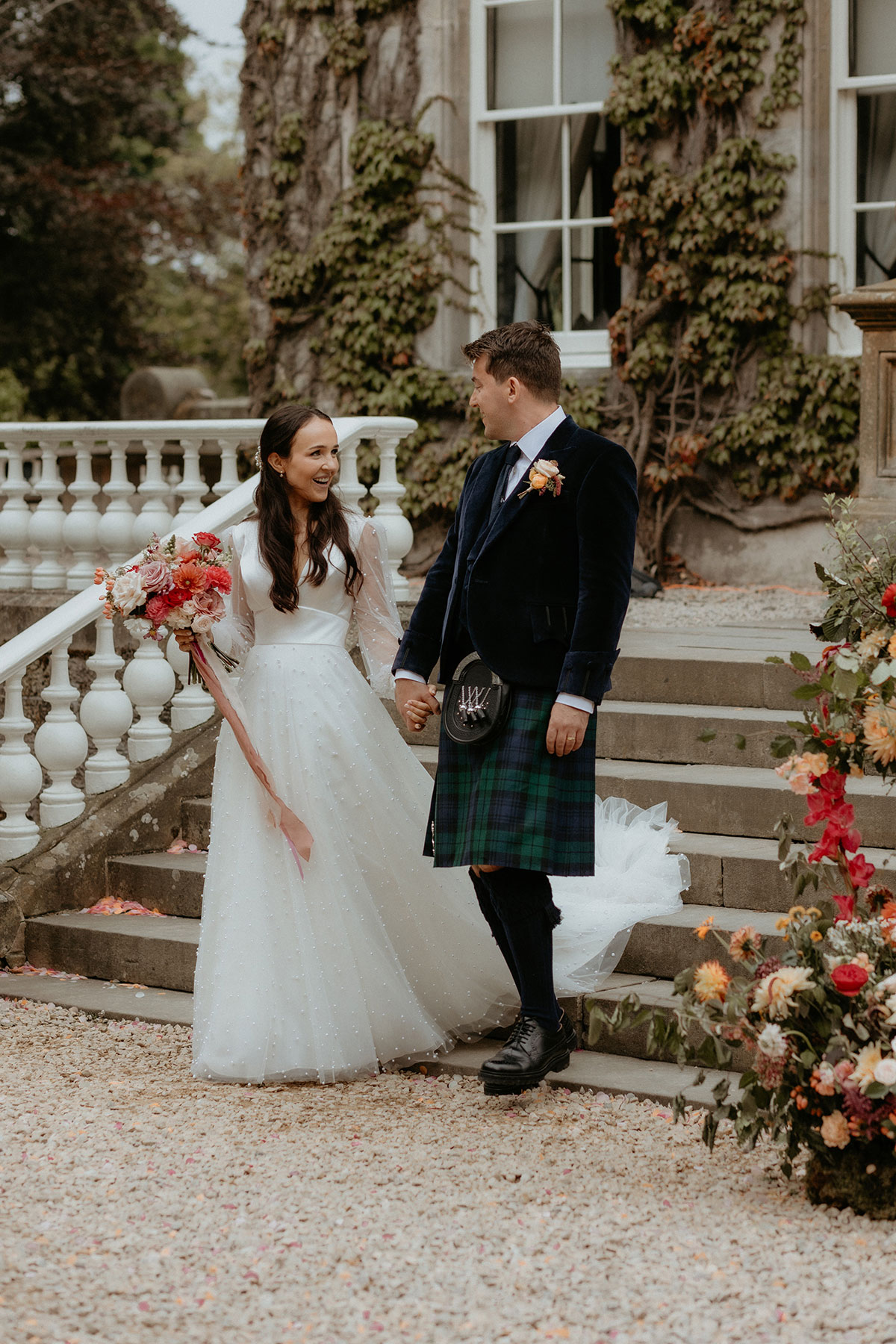 a smiling bride and groom in a kilt walking down a set of stone stairs outside Carlowrie Castle