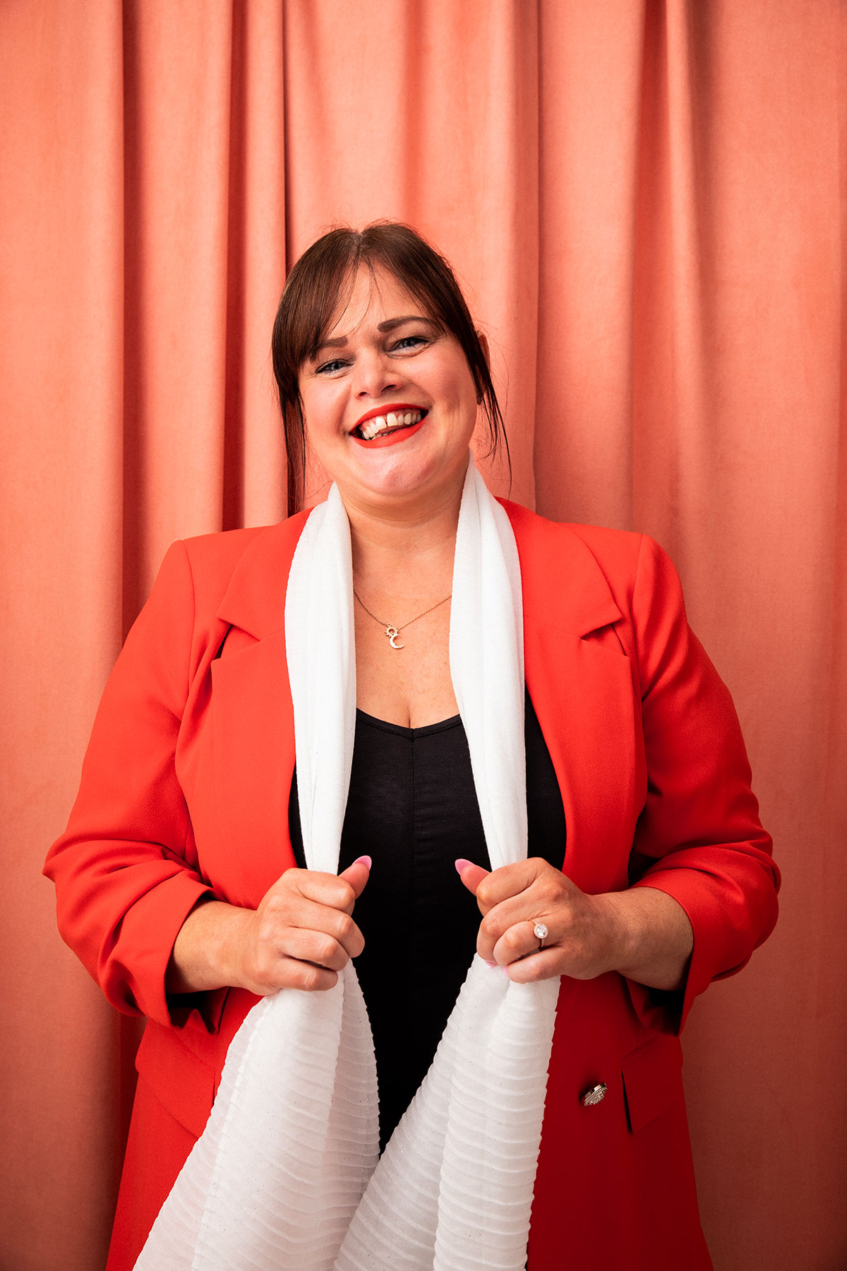 Portrait of Sharon Mullen in red blazer and white scarf against coral backdrop, Scottish wedding supplier headshot
