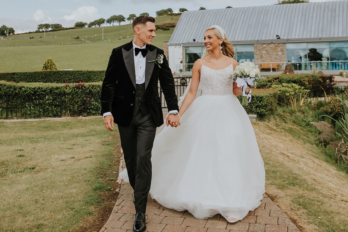 blonde bride in ballgown holding bouquet in one hand and groom in tux in the other, walk down brick path with grassy hills behind them
