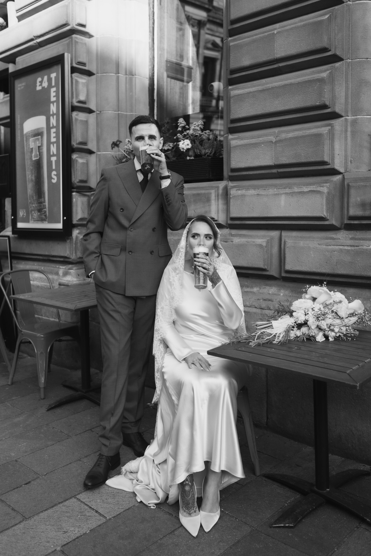 Black-and-white portrait of the bride and groom enjoying pints outside a Glasgow pub on their wedding day.