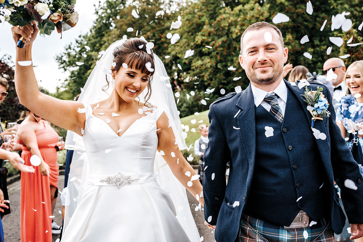 bride and groom walk towards camera smiling while their guests throw white coloured confetti on them