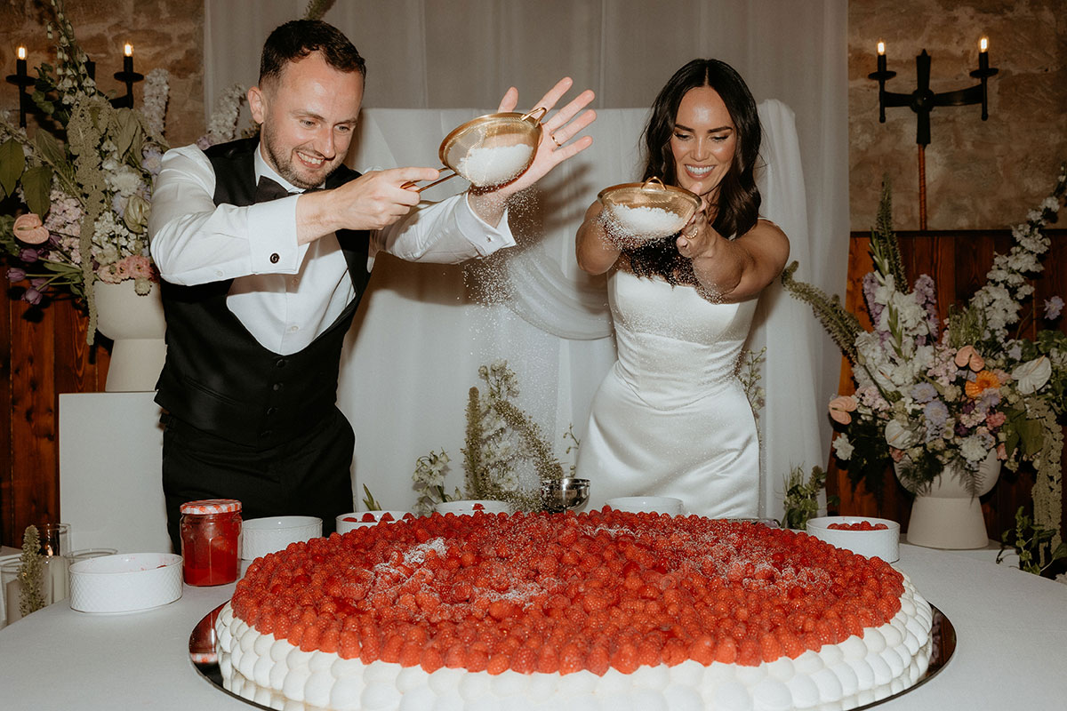 Bride and groom dusting icing sugar over giant pavlova wedding cake at Rosebery Steading reception in Midlothian.