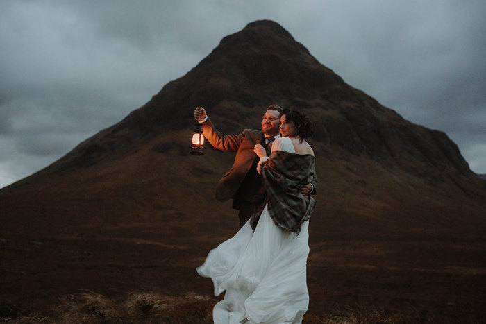 a groom holding a lantern at twilight while posing with a bride against a mountain in Glencoe