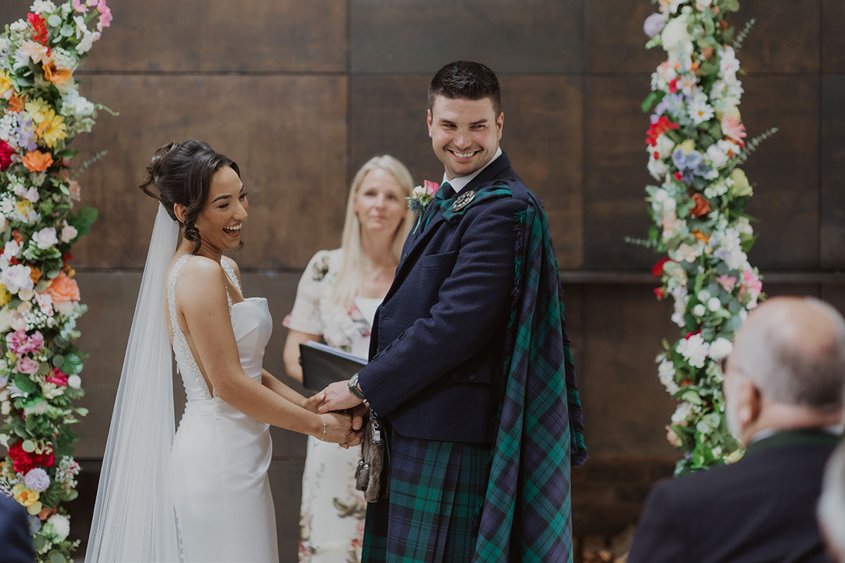 Bride and groom smiling during indoor Scottish wedding ceremony, groom wearing traditional tartan kilt beneath floral arch