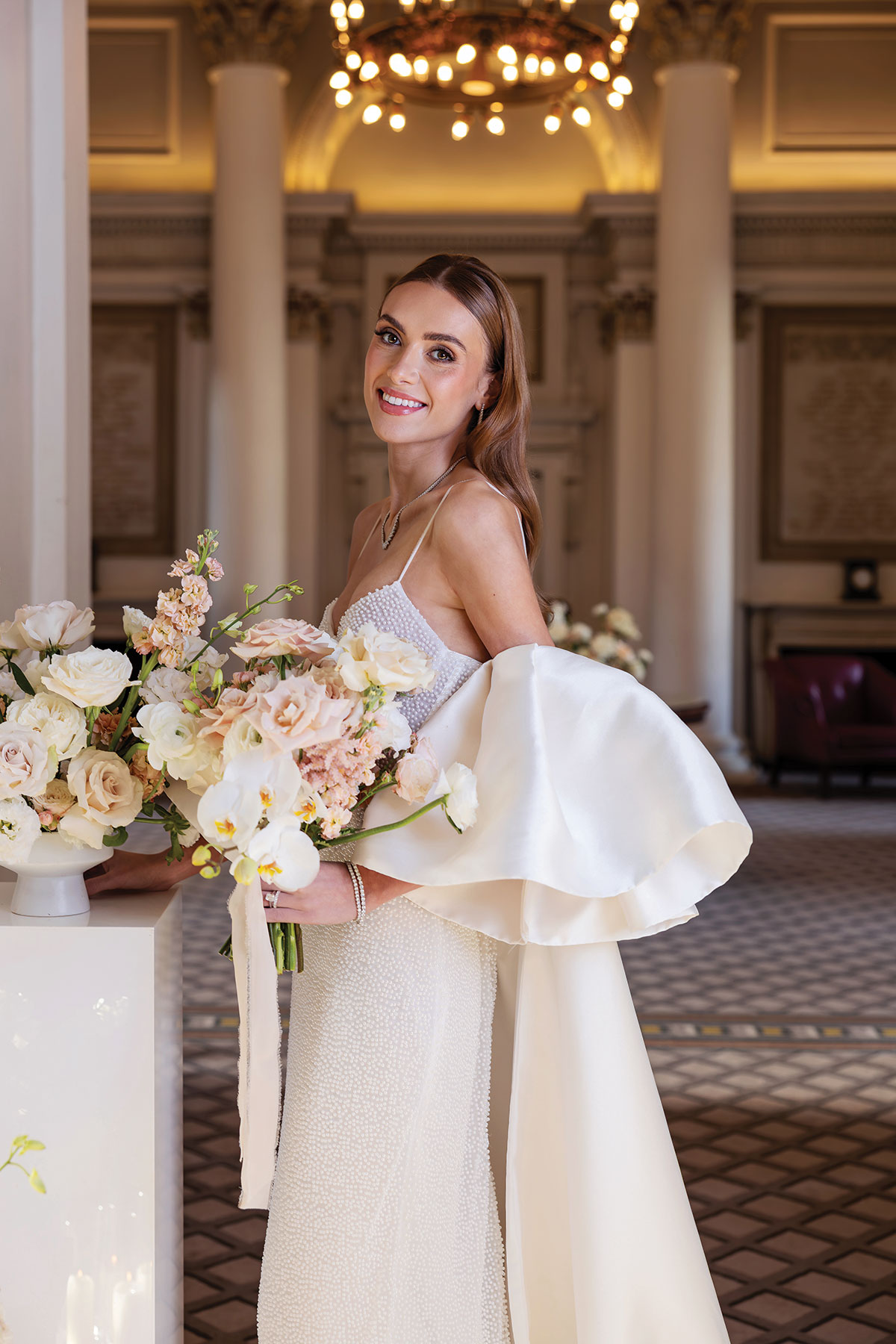 Bride holding peach and ivory bouquet in grand historic Edinburgh venue with chandeliers and columns