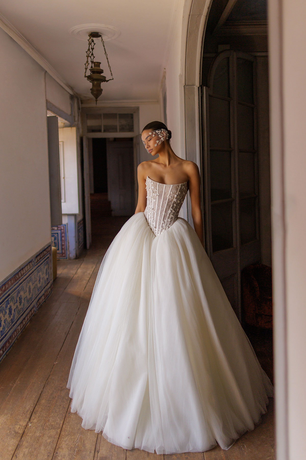 Bride wearing a corset ballgown wedding dress with structured bodice, voluminous tulle skirt and short veil, posed in a softly lit historic hallway with wooden floors