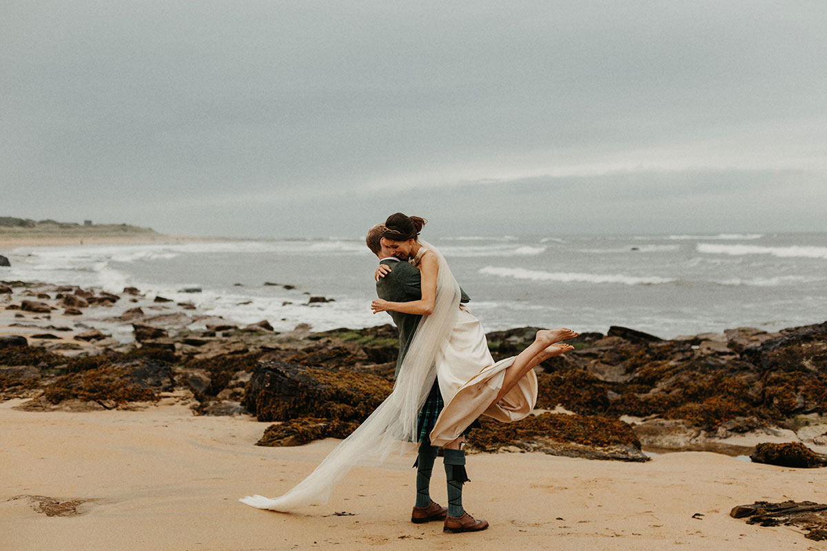 Bride and groom embracing on a windswept beach at Cambo Estate, with the groom lifting the bride as her long veil trails behind them against the rocky shoreline