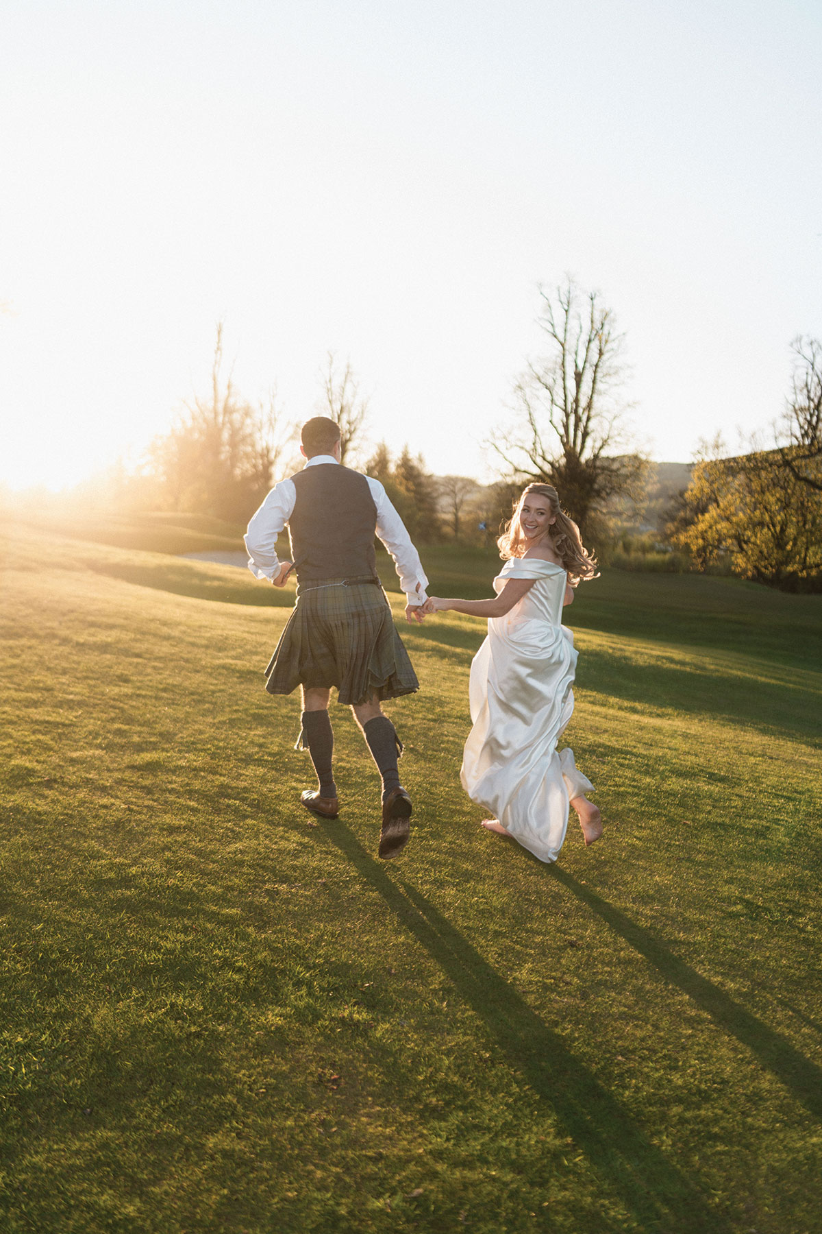 Bride and groom running hand in hand across a Scottish golf course at golden hour after their wedding ceremony.