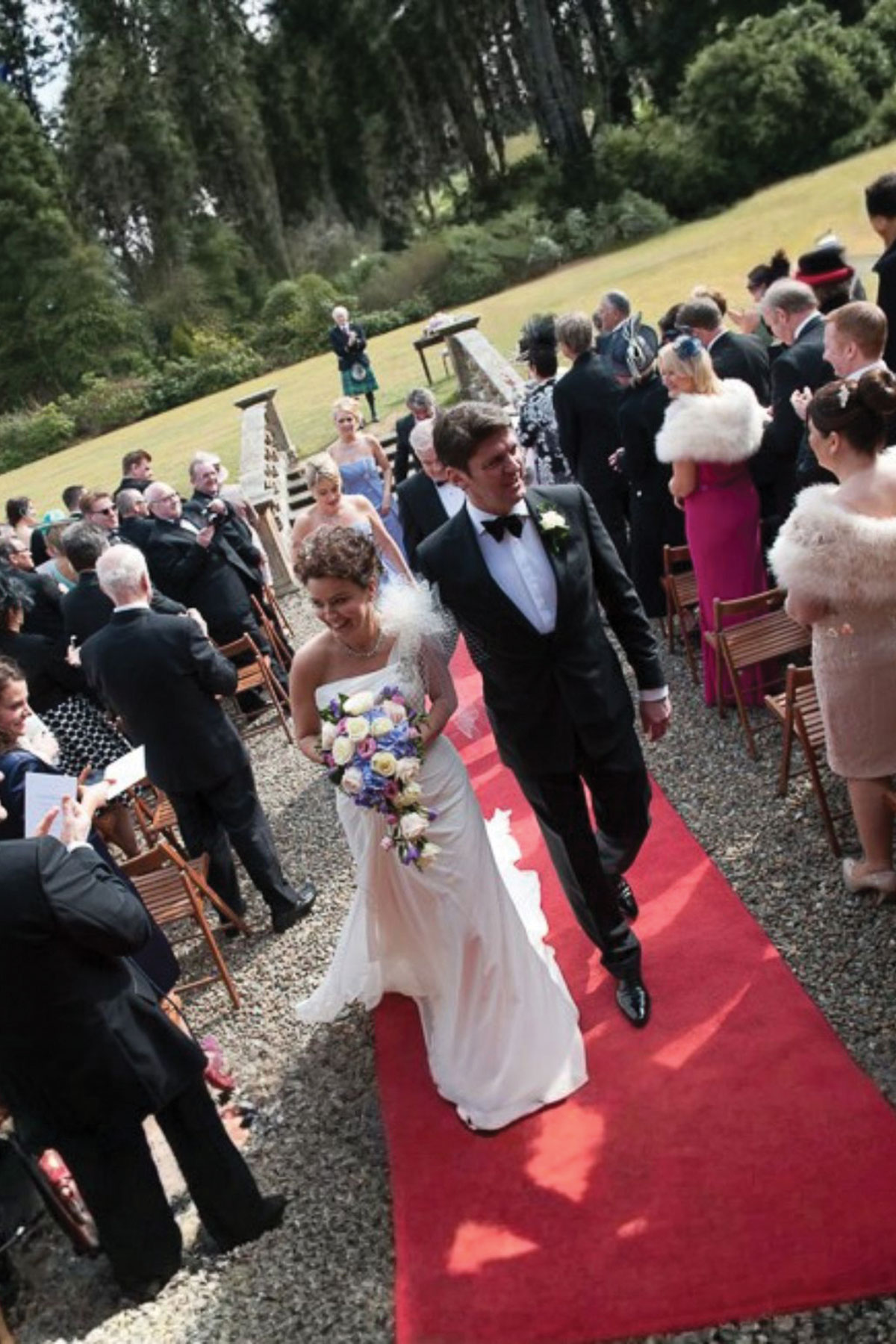 Bride and groom walking down a red carpet aisle outdoors after their ceremony, surrounded by seated guests in formal attire