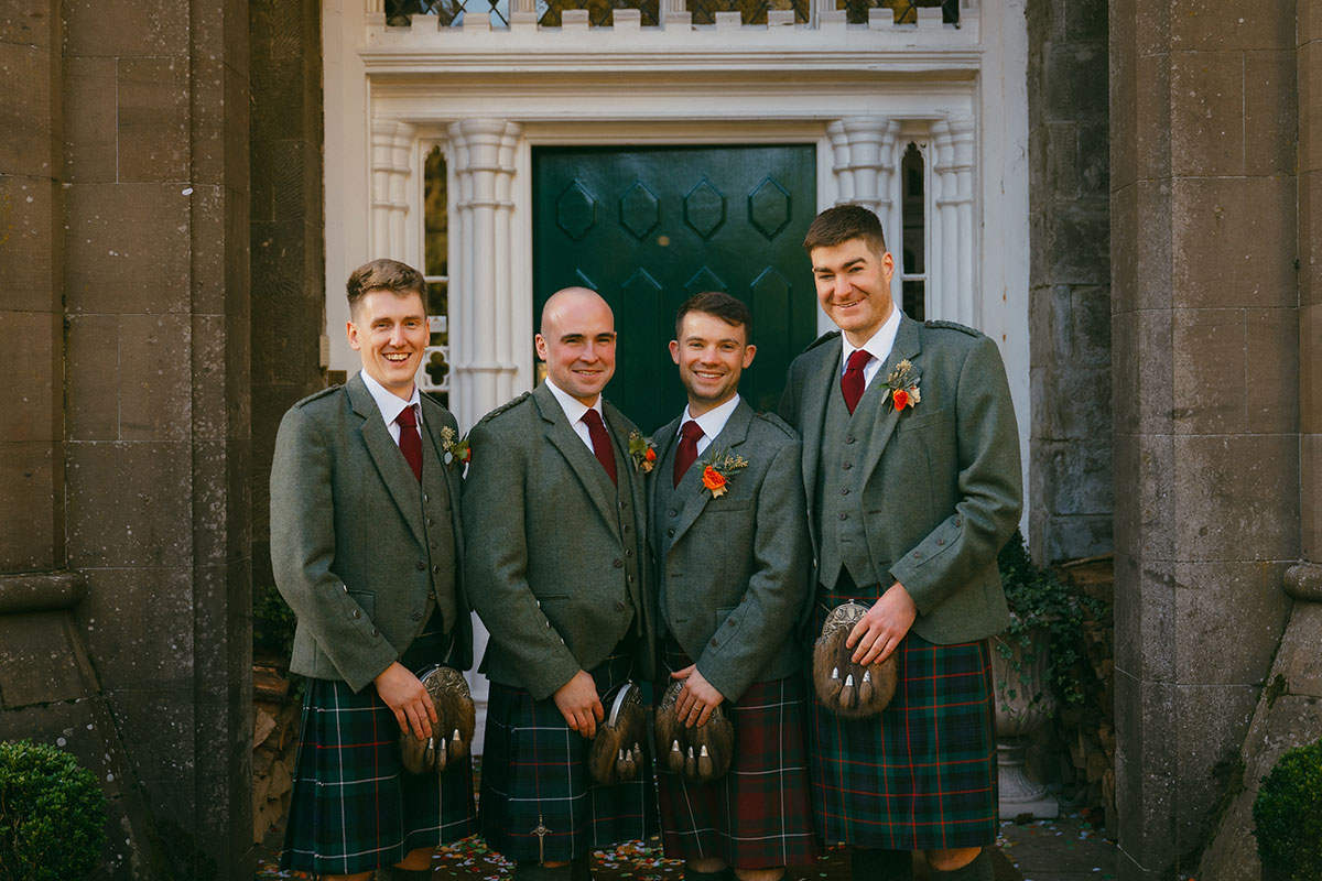 Groom and groomsmen in traditional Scottish kilts posing outside castle wedding venue entrance