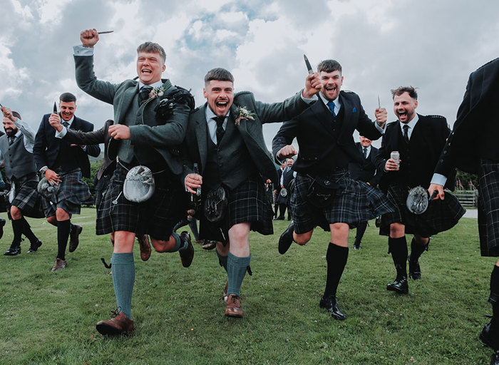 men in kilts at a wedding running over grass towards the camera while holding their sgian dubhs in the air