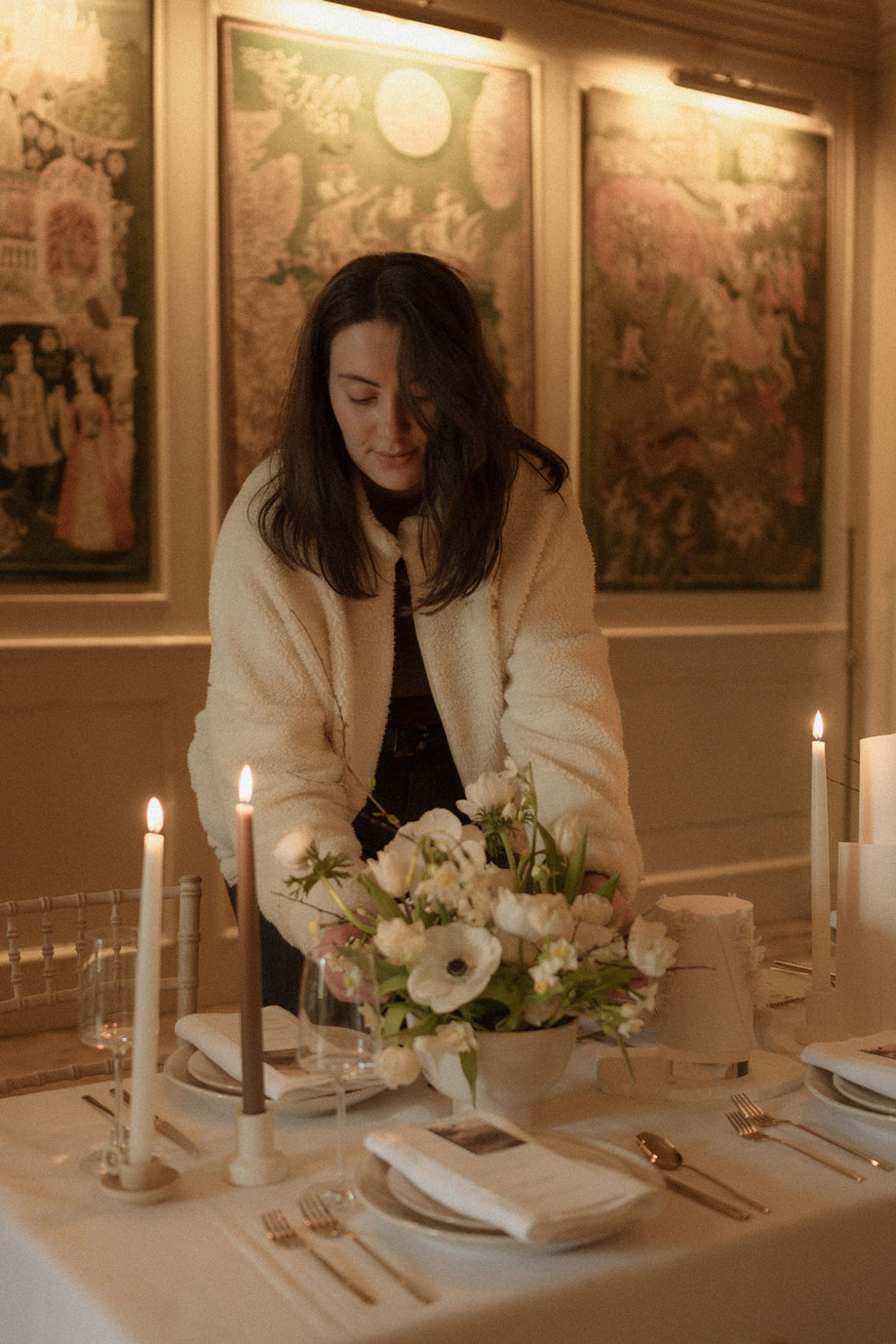 brunette woman stands in dimmed light, arranging a vase of neutral flowers on a tablecloth set dining table, complete with candlesticks, wine glasses, and multiple place settings