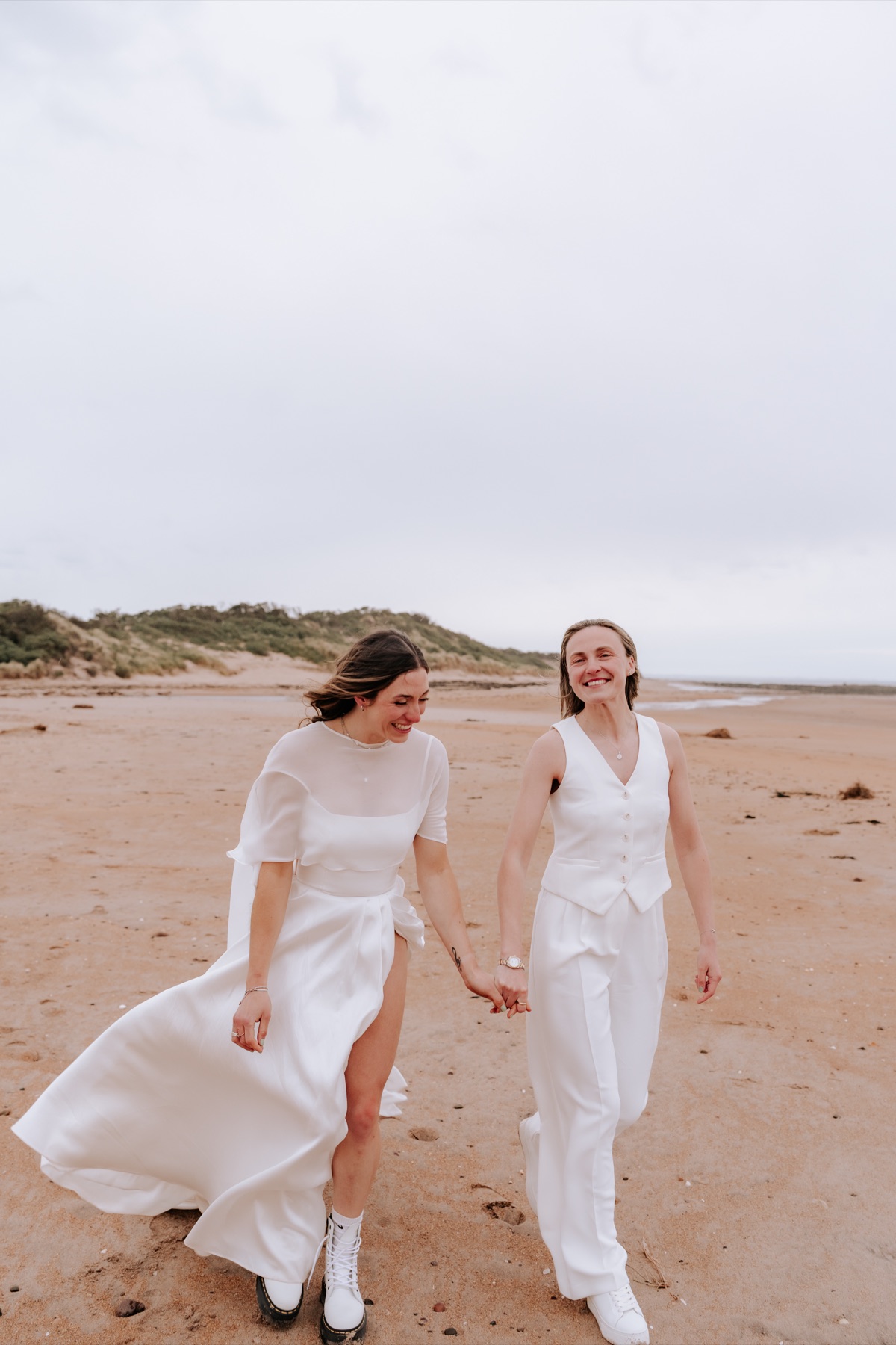 Hannah and Donna walking hand in hand along the beach in their wedding outfits.
