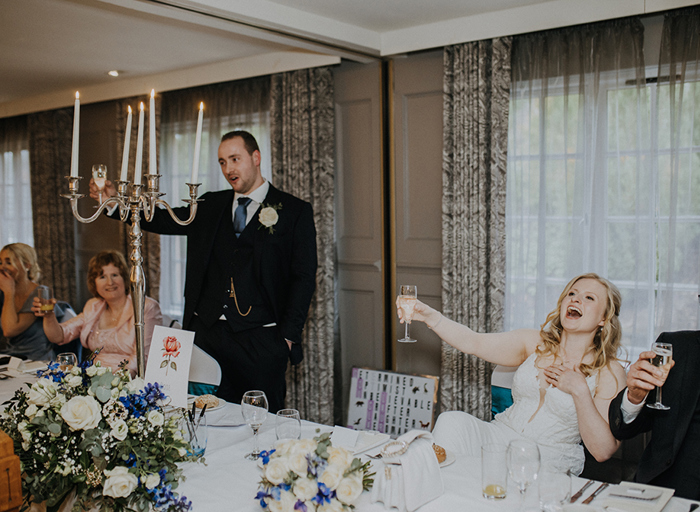 a bride laughing as she raises a glass of champagne as the man next to her gives a speech also holding up a glass of champagne