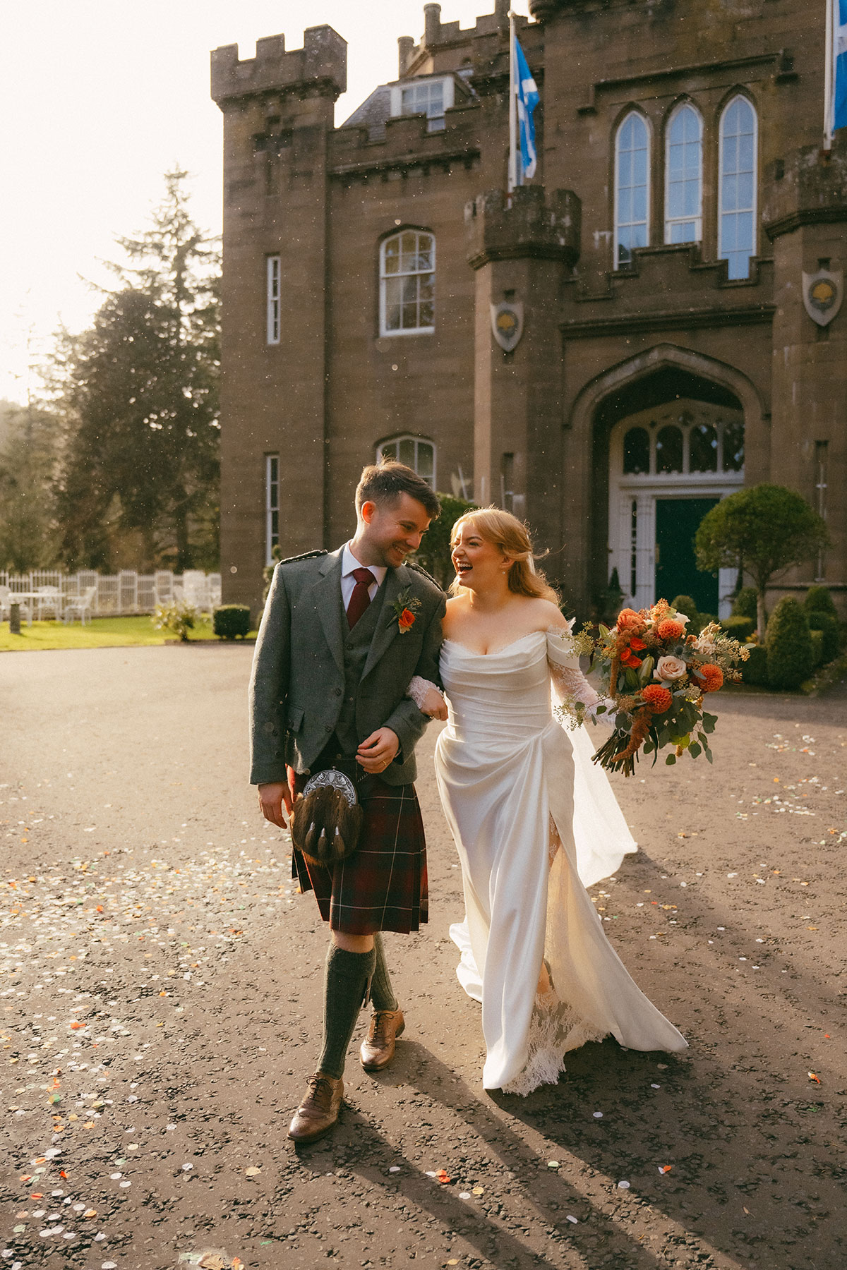 Bride and groom walking together outside Scottish castle wedding venue with confetti on ground and autumn bouquet
