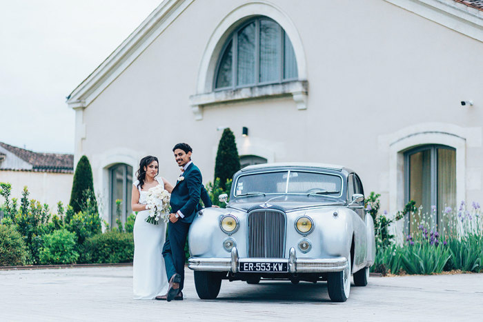 A bride and groom posing for a picture next to a car outside Chais Monnet & Spa in Cognac