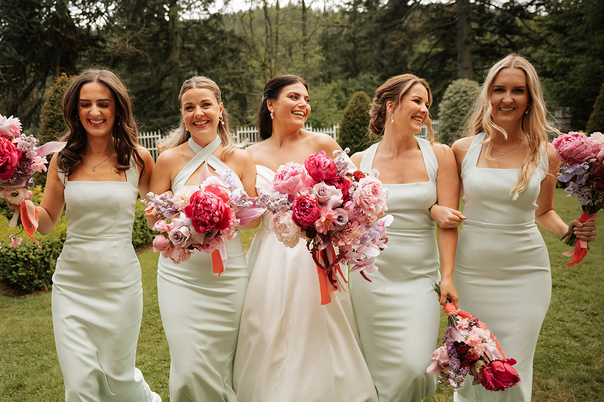 Bride and bridesmaids walking in garden at Drumtochty Castle Aberdeenshire holding pink and red wedding bouquets