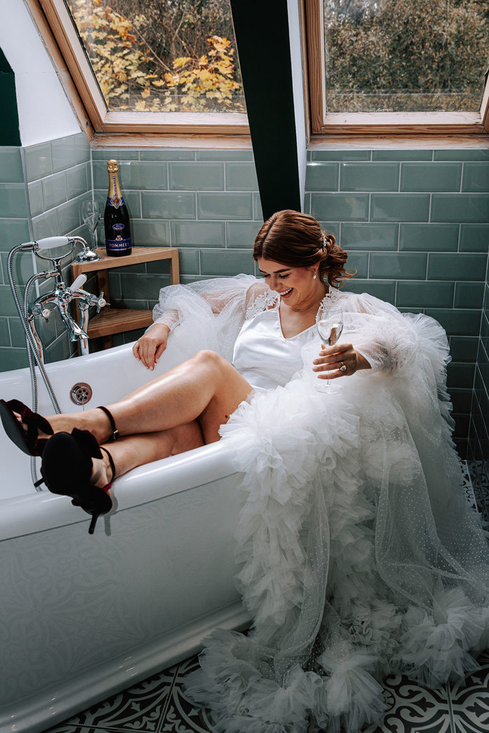 brunette bride sits in an empty bath wearing a white silk slip, frilly tulle robe and black heels while holding a glass of champagne