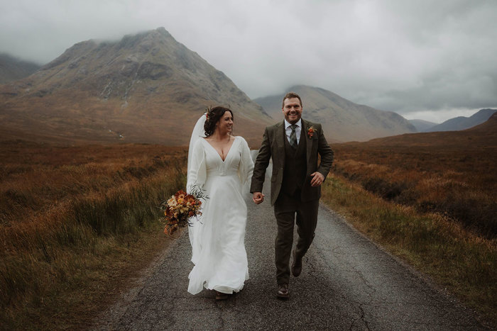 a bride and groom walking along a narrow road in the shadow of Buachaille Etive Mòr