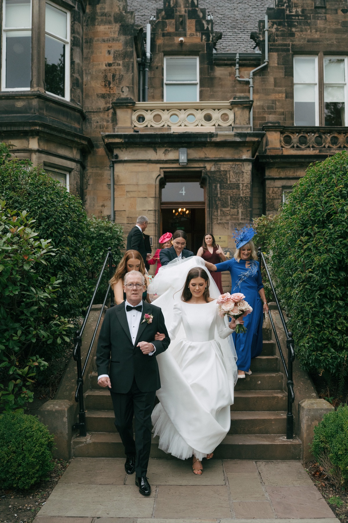 Bride Nicole walks down the steps of her wedding preparation venue with her dad, carrying a pink rose bouquet as guests follow behind.