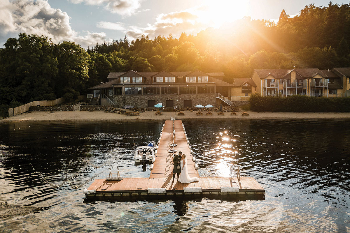 bride and groom stand side by side out on the end of a wooden dock, with stone building in background and sun setting overhead