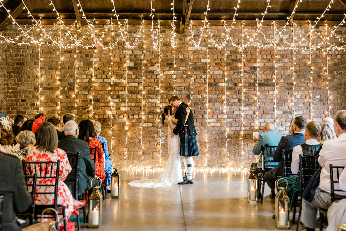 Bride and groom sharing their first kiss during an indoor wedding ceremony beneath fairy lights
