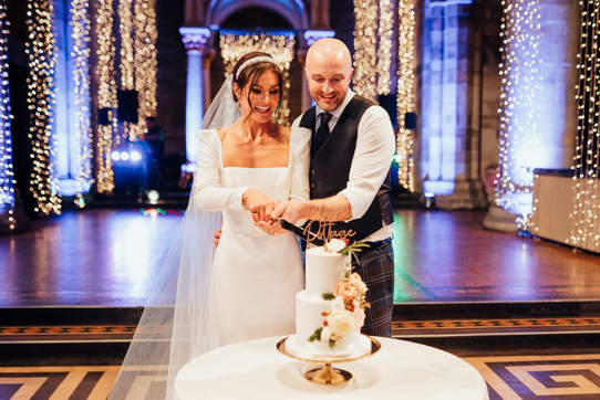 couple cutting wedding cake smiling on dancefloor in front of twinkling fairy lights