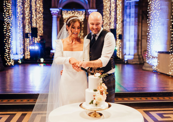 couple cutting wedding cake smiling on dancefloor in front of twinkling fairy lights