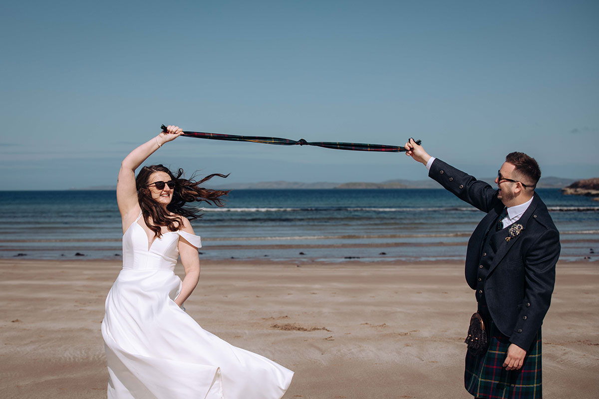 Bride and groom laugh as they hold their tartan ribbon during handfasting on Achnahaird Bay beach in the Scottish Highlands.