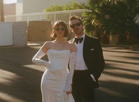 A couple dressed in wedding attire stands together outdoors, with palm trees and a building in the background