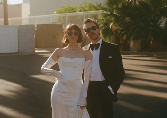 A couple dressed in wedding attire stands together outdoors, with palm trees and a building in the background