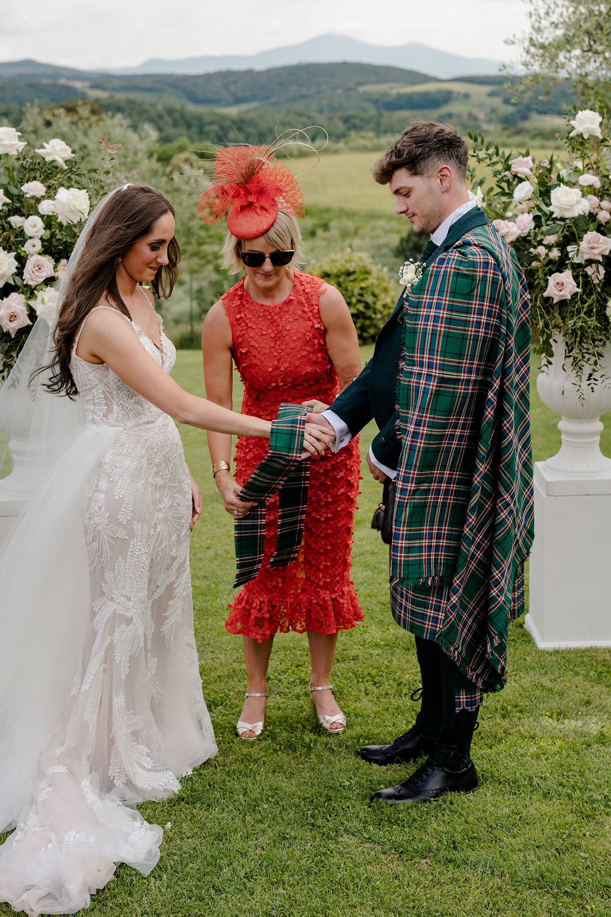blonde woman with bob leads bride and groom in tying the knot with tartan cloth, wearing a red-orange dress with 3D flowers and a matching percher hat decorated with veiling, quills and feather flowers