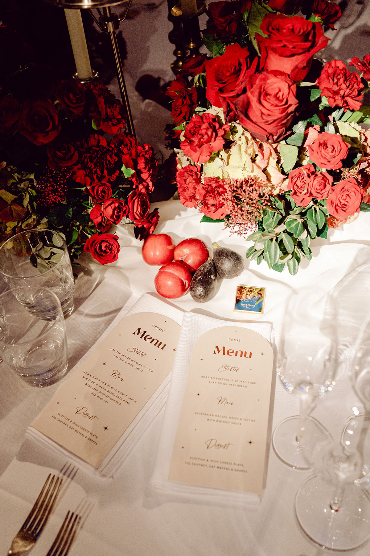 Close-up of wedding table décor with deep red florals, candles and printed menus laid out for the bride and groom