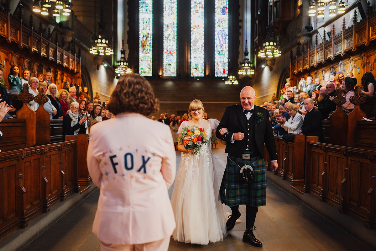 Bride walking up the aisle on her father’s arm toward her partner, with guests standing in the wooden pews and stained-glass windows illuminating the chapel.