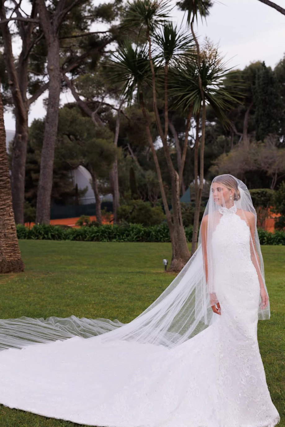 bride in halterneck wedding dress and long train stands with veil covering her face and upper body while the rest of the veil flows along the back of the dress