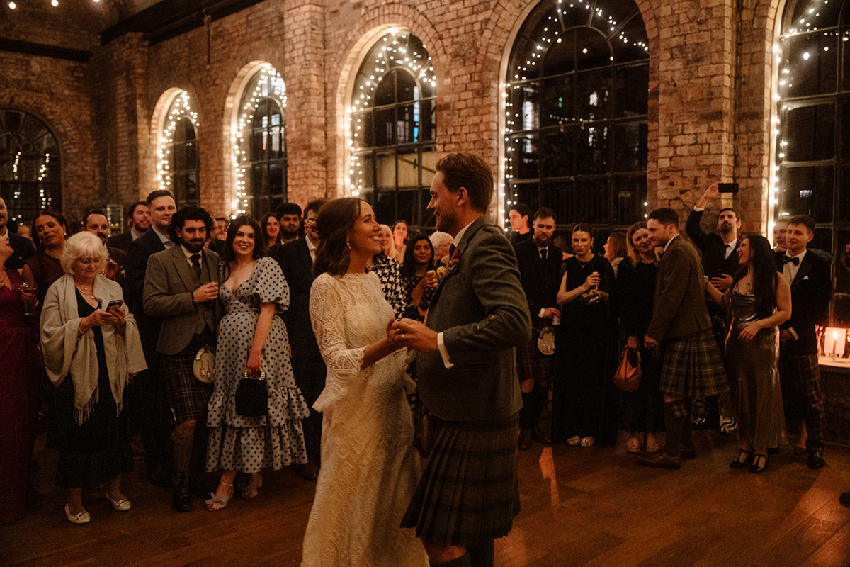 Newlyweds share their first dance surrounded by wedding guests in a historic industrial wedding venue decorated with fairy lights and exposed brickwork