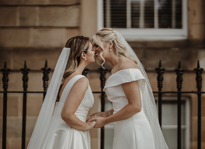 Two brides kissing with black railings and building in the background