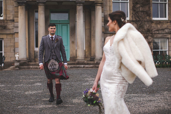 A groom in a grey and red kilt walking towards a bride in a fitted white dress with a white fur coat and a bouquet of flowers